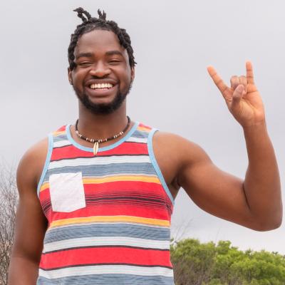 A smiling young man wearing a colorful striped tank top, standing outdoors and making the “Hook ’em Horns” hand sign, a symbol associated with the University of Texas at Austin. The image conveys pride and enthusiasm for the PACE co-enrollment program with UT Austin.